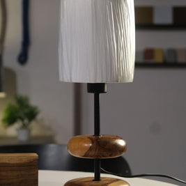 A serene forest retreat bedside features the 'Earthy Wood Glow' table lamp—teak wood base, sleek metal stem, and off-white fabric shade—on a rustic wooden table, with a golden glow illuminating books, a ceramic mug, and a lush forest view through a window