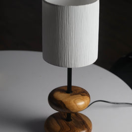 A warm teak wood table lamp with a sleek metal stem and soft off-white fabric shade glows gently on a rustic wooden bedside table in a forest retreat bedroom, surrounded by nature-inspired decor and a view of pine trees through a window.