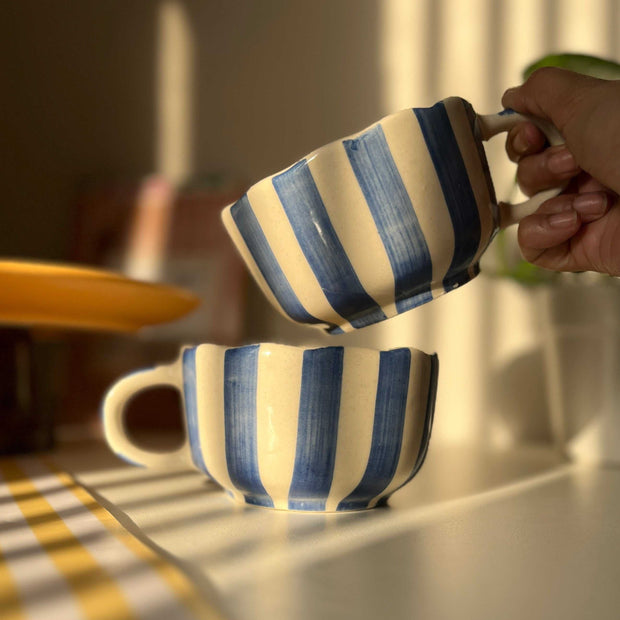 Hand holding a blue striped mug among various colorful mugs on a wooden surface. caprice striped pattern. 