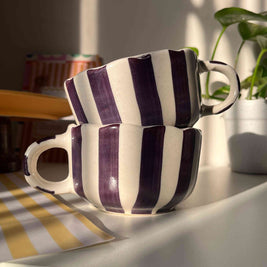 Pair of striped purple coffee and soup mugs stacked in a set of two on a hook with a plant in the background