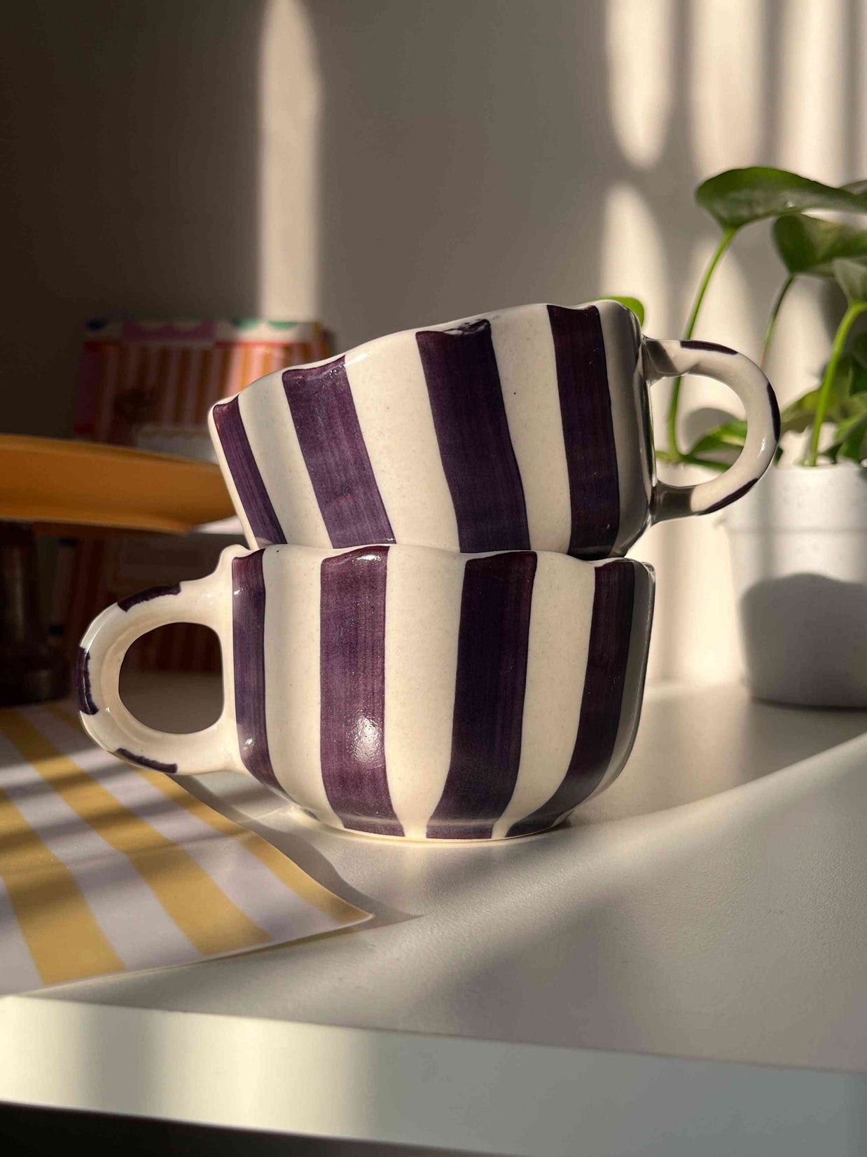 Pair of striped purple coffee and soup mugs stacked in a set of two on a hook with a plant in the background