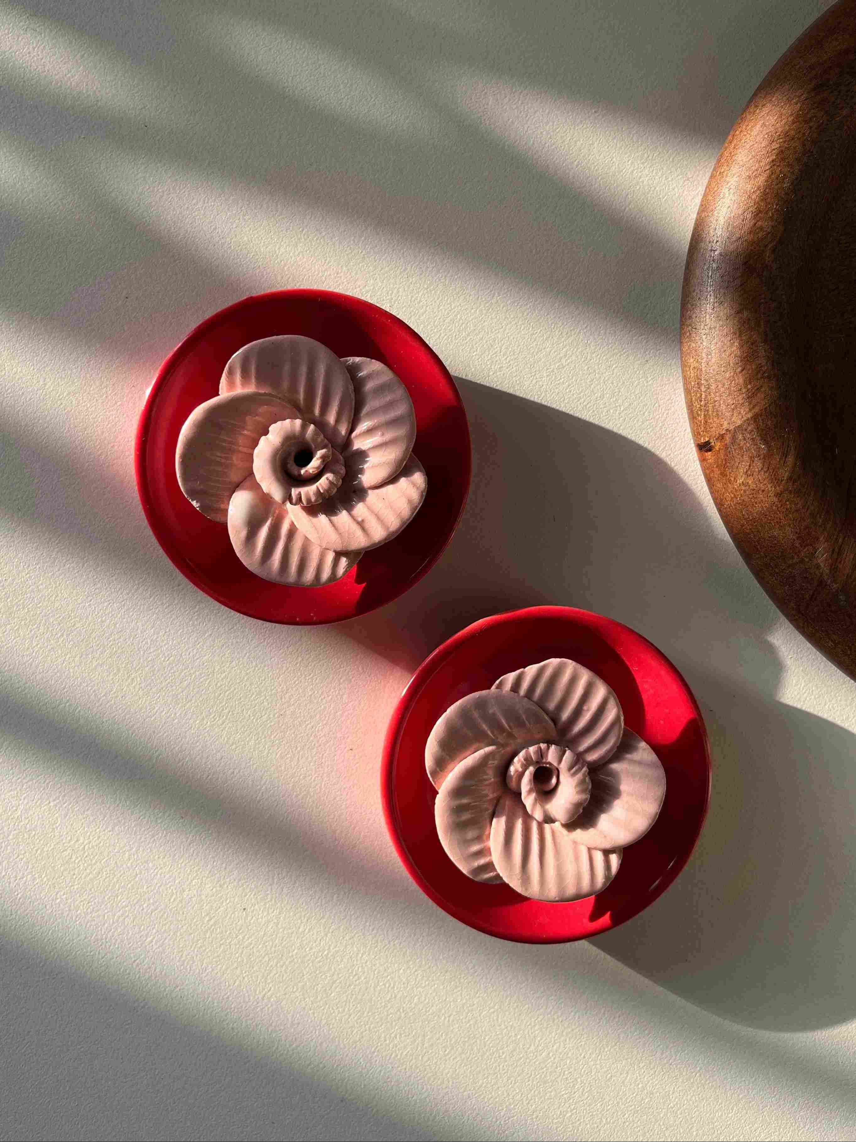 Two red ceramic flower-shaped objects on a white surface with shadows. They hold incense sticks. 