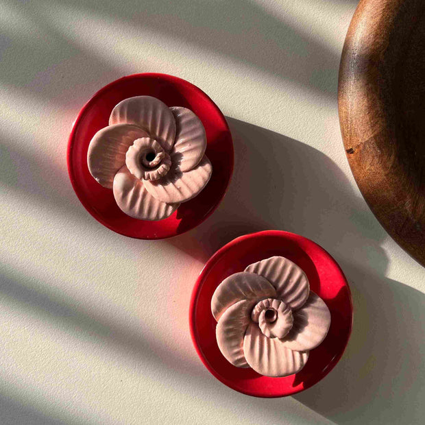 Two red ceramic flower-shaped objects on a white surface with shadows. They hold incense sticks. 