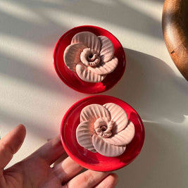 Two red containers with shell-like designs on a white surface, one of which is held by a hand.