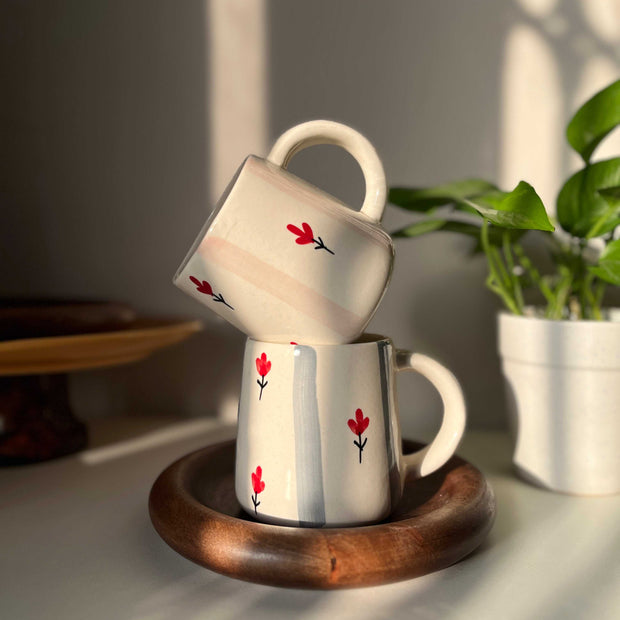 Two ceramic mugs with red flower designs on a wooden coaster, placed on a surface with a plant in the background.
