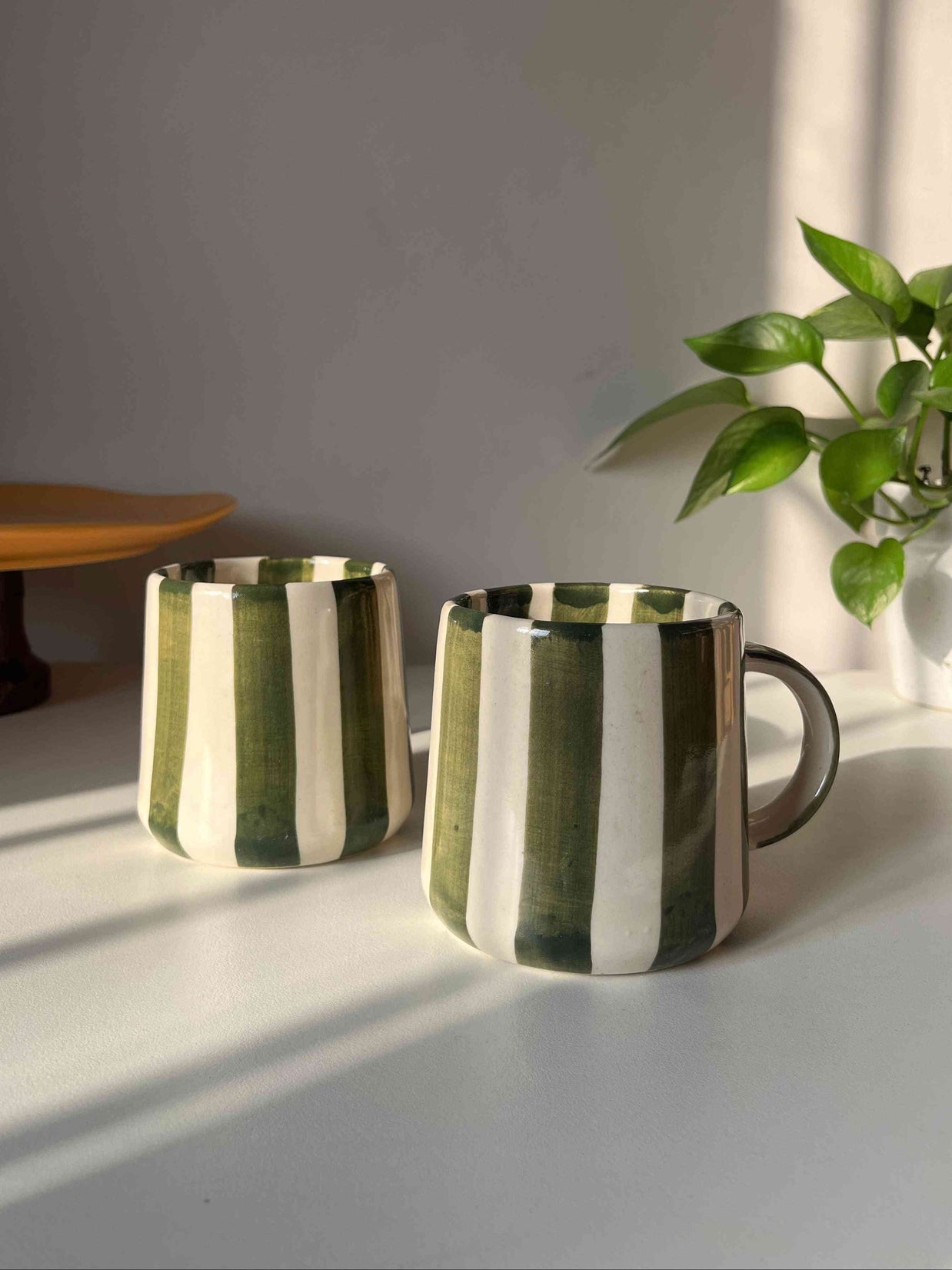 Two green striped ceramic coffee mugs on a light surface with a plant in the background