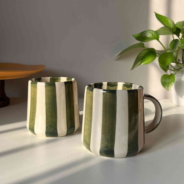 Two green striped ceramic coffee mugs on a light surface with a plant in the background
