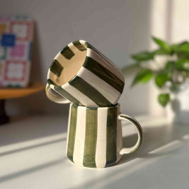 Two ceramic mugs stacked on a light surface with a plant and colorful box in the background.