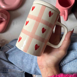 Hand holding a mug with red heart pattern on a checkered background