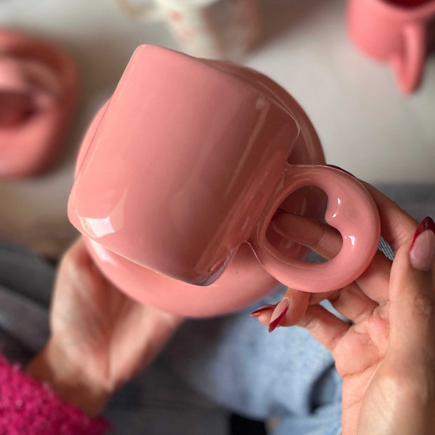 Pink ceramic cup with a round saucer. The handle of the mug is heart shaped. 