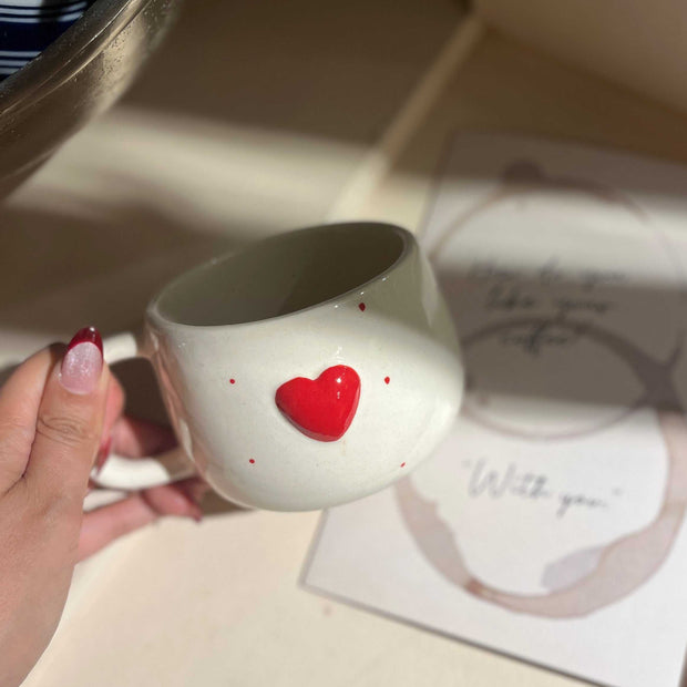 Person holding a white mug with a red heart design on a surface with a card and decorative item.
