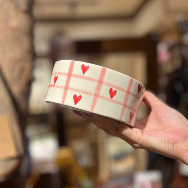 Ceramic cup with red heart pattern held by a hand in a blurred indoor setting