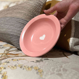 Hand holding a pink ceramic bowl with a heart design on a textured surface.