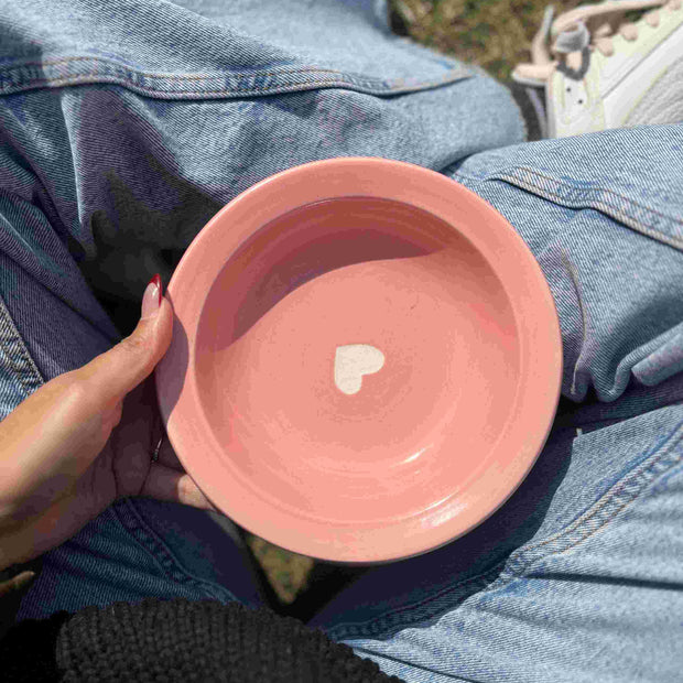 Pink ceramic bowl with a heart design held by a person wearing blue jeans.