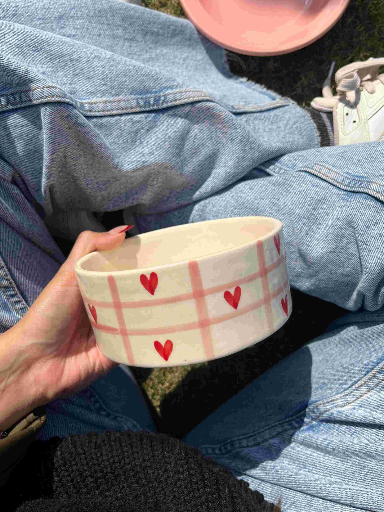 Person holding a ceramic bowl with heart patterns, sitting on grass. checkered pink with red hearts