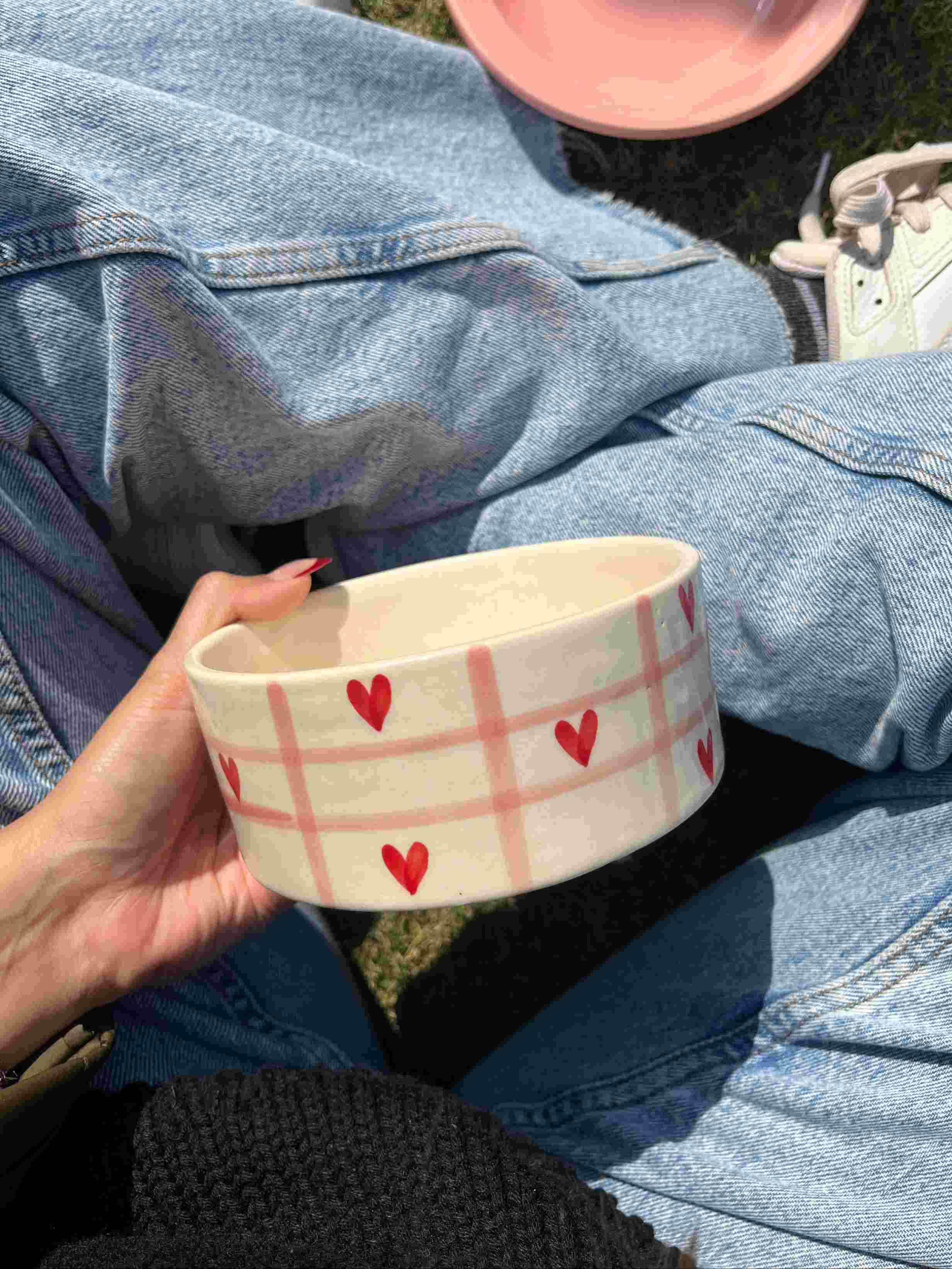 Person holding a ceramic bowl with heart patterns, sitting on grass. checkered pink with red hearts