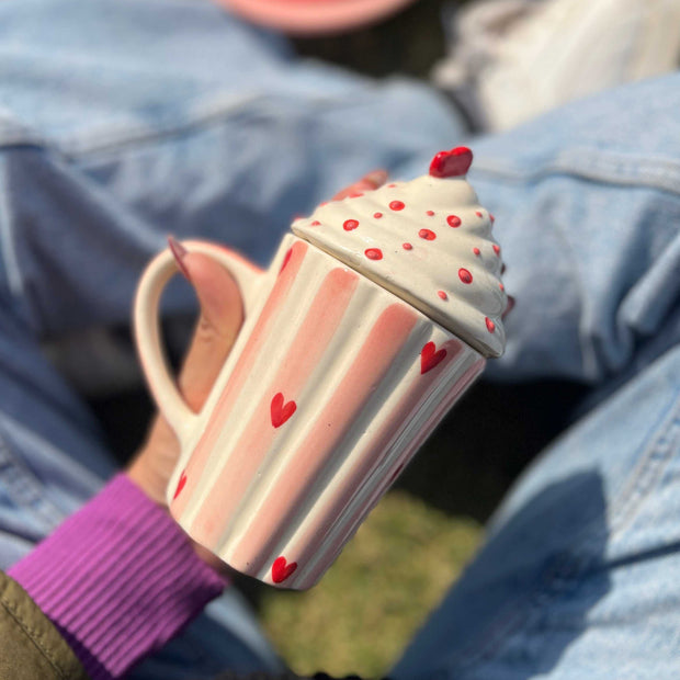 Cupcake-shaped mug with pink handle held by a person wearing a purple sleeve, sitting on blue jeans.