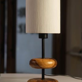 On a honey-toned wooden bedside table in a forest retreat, the 'Earthy Wood Glow' lamp with its teak wood base, metal stem, and off-white shade casts a soft light, framed by a moss-green wall, nature sketches, and a view of tall pine trees outside.