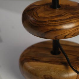The 'Earthy Wood Glow' lamp, with its earthy teak wood base, minimalist metal stem, and soft off-white shade, rests on a wooden bedside table in a tranquil forest retreat, surrounded by natural decor and a scenic view of pine trees and dappled sunlight.