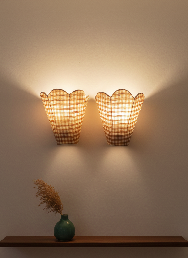 Two wall-mounted lamps with checkered shades on a plain wall, with a green vase and dried plants on a shelf below.