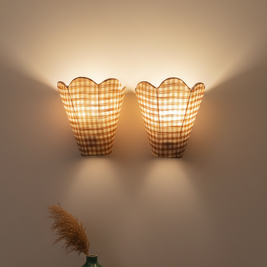 Two wall-mounted lamps with checkered shades on a plain wall, with a green vase and dried plants on a shelf below.