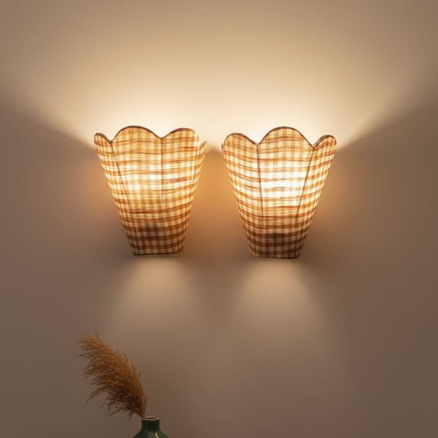 Two wall-mounted lamps with checkered shades on a plain wall, with a green vase and dried plants on a shelf below.