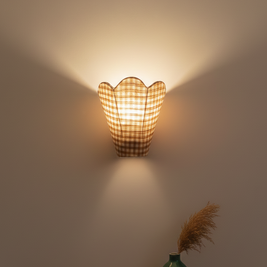 Wall-mounted lamp with a checkered pattern casting light on a plain wall, with a vase and dried plants on a shelf below.