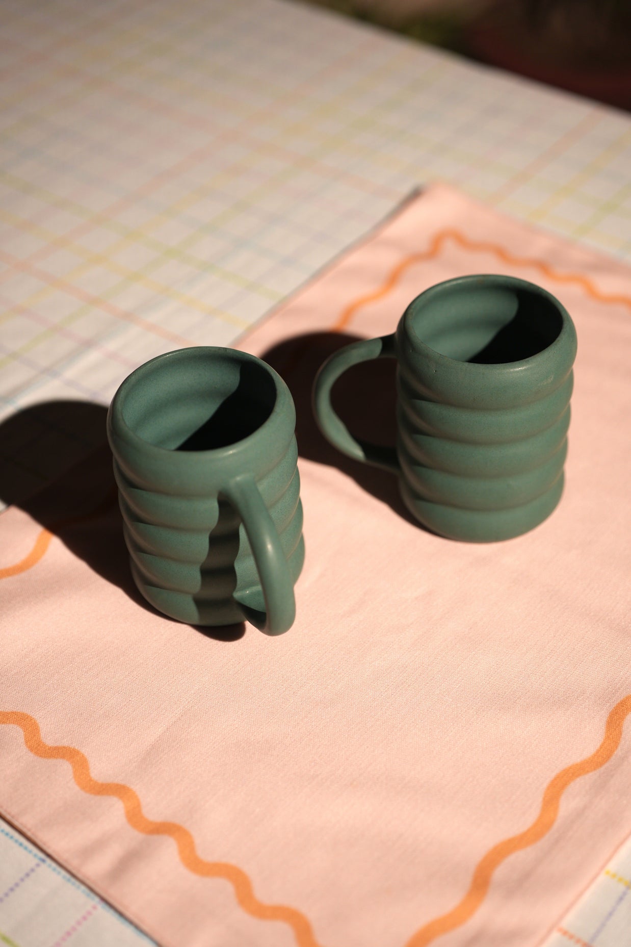 Two green ceramic mugs on a pink cloth with a grid background