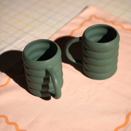 Two green ceramic mugs on a pink cloth with a grid background