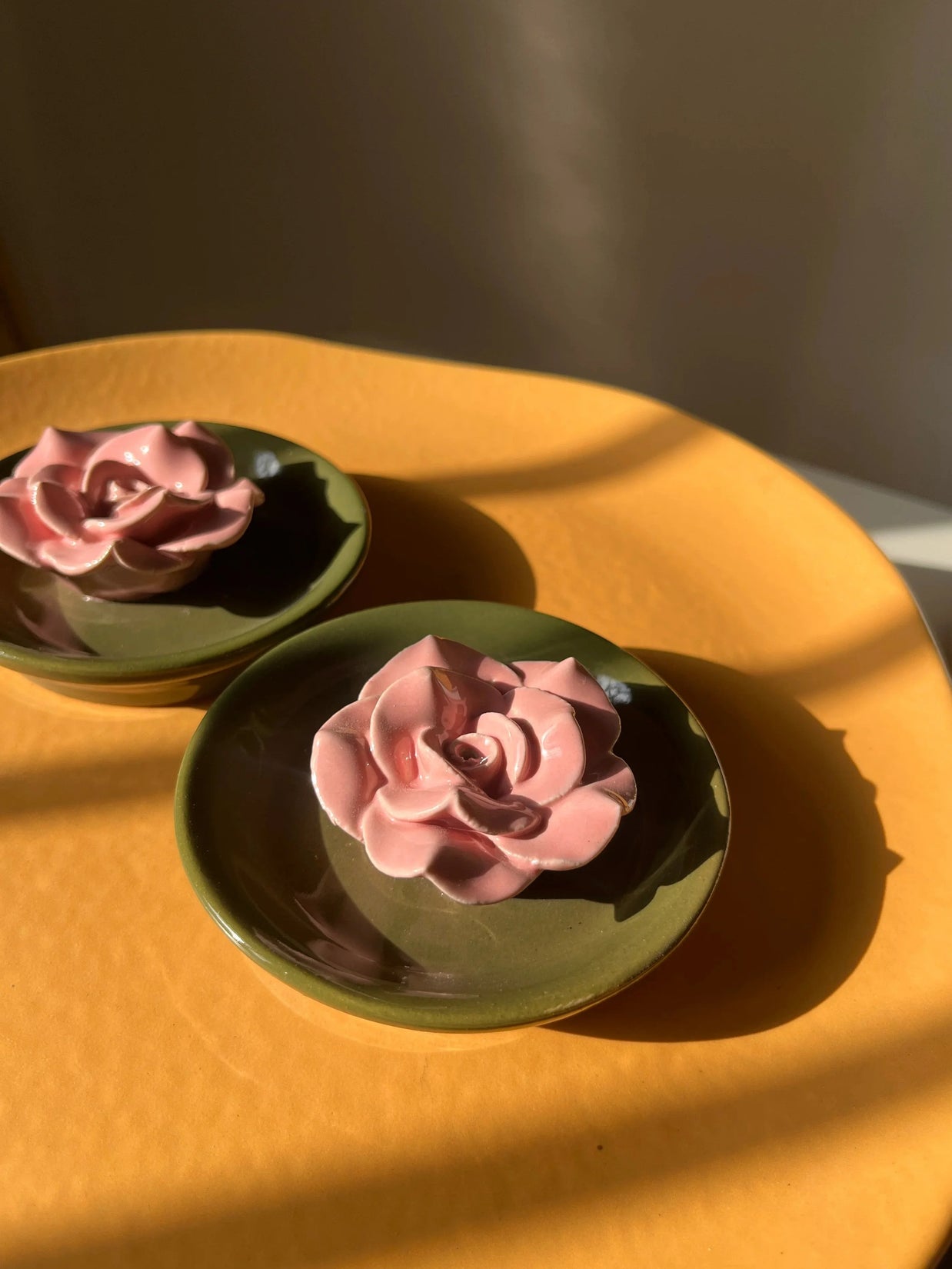 Two pink rose-shaped incense stick holders, placed on a warm yellow-orange surface with strong sunlight and shadows.