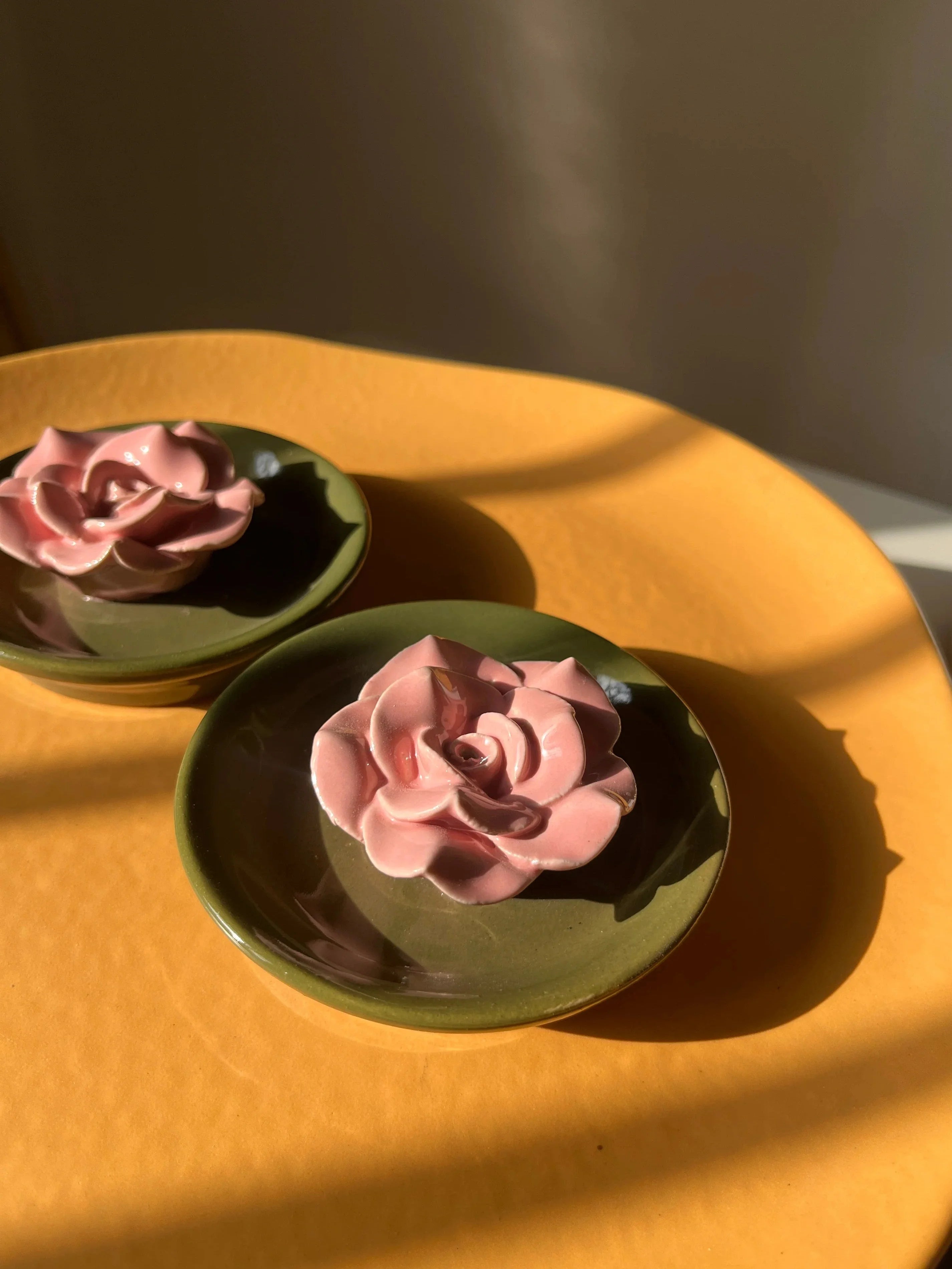 Two pink rose-shaped incense stick holders, placed on a warm yellow-orange surface with strong sunlight and shadows.