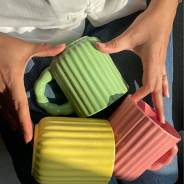 Three colorful mugs (green, yellow, pink) held by a person with a white shirt and blue jeans.