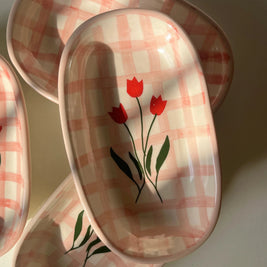 A set of oval ceramic dishes with a pink and white gingham check pattern and a red tulip design, arranged on a light surface with strong natural shadows.