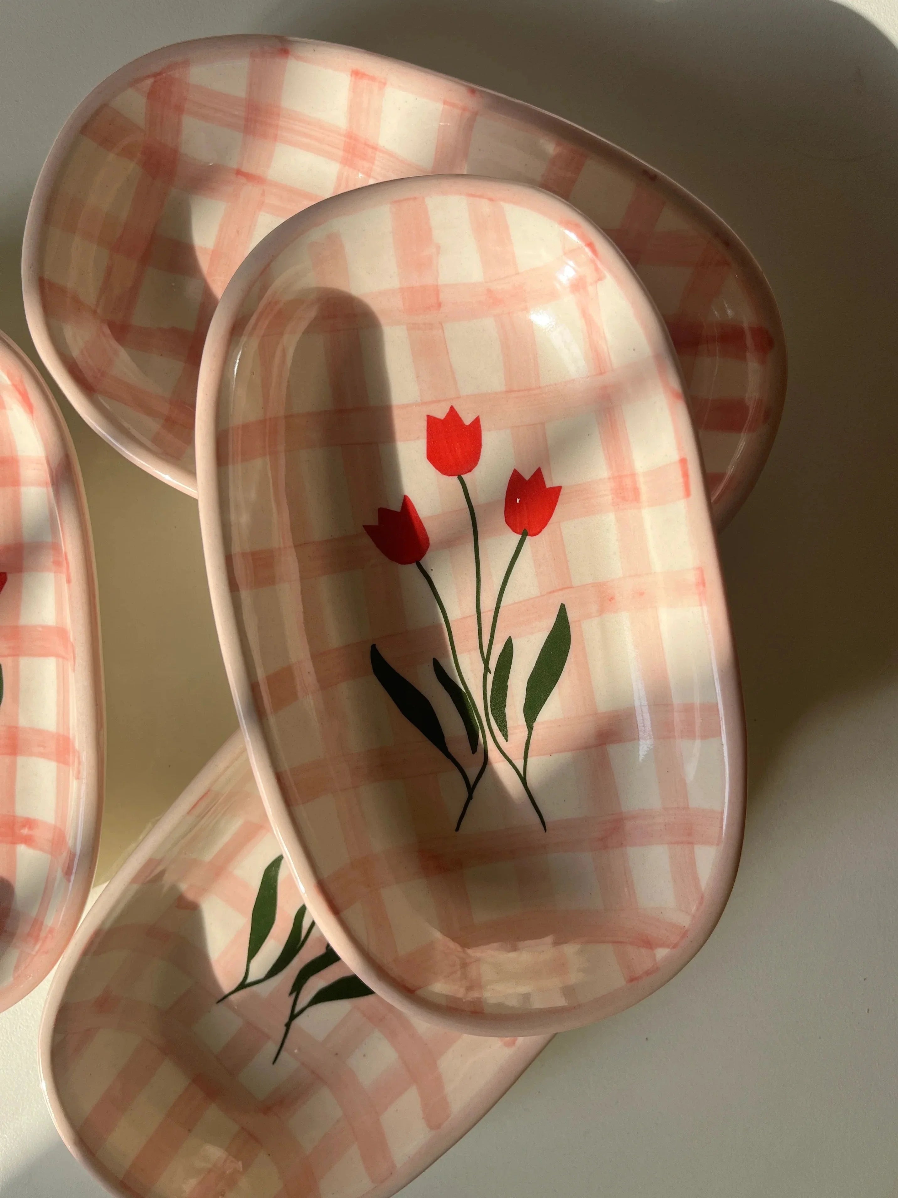 A set of oval ceramic dishes with a pink and white gingham check pattern and a red tulip design, arranged on a light surface with strong natural shadows.
