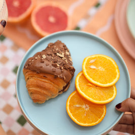 Person holding a blue ceramic pastel quarter plate with a croissant and orange slices on a colorful tablecloth.