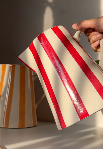 Red and white striped ceramic pitcher held by a hand with another pitcher in the background.