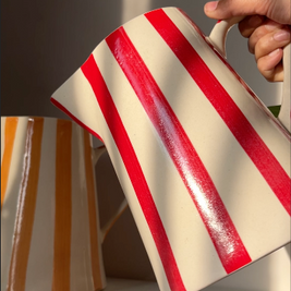 Red and white striped ceramic pitcher held by a hand with another pitcher in the background.