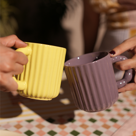 Two hands holding a yellow and a purple textured mug on a patterned tablecloth.