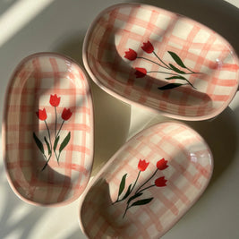 Three oval ceramic dishes with a pink and white plaid pattern and red tulip designs, arranged on a white surface with natural window shadows.