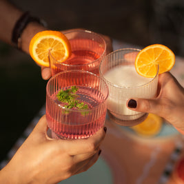 Three glasses of pink and clear drinks with orange slices held by people against a blurred background.