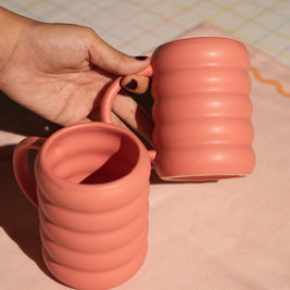 Hand holding two pink-colored ceramic tea and coffee mugs on a patterned surface