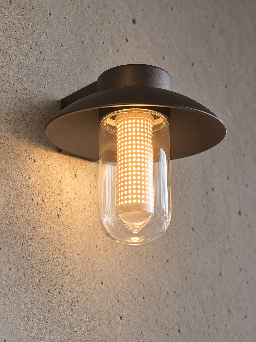A modern outdoor wall sconce with a dark bronze metal frame and a clear glass cylinder, glowing with a warm yellow light against a textured concrete wall.