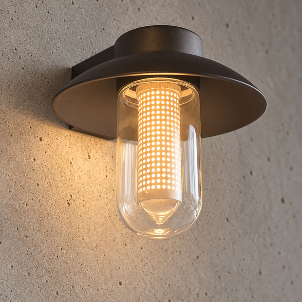 A modern outdoor wall sconce with a dark bronze metal frame and a clear glass cylinder, glowing with a warm yellow light against a textured concrete wall.