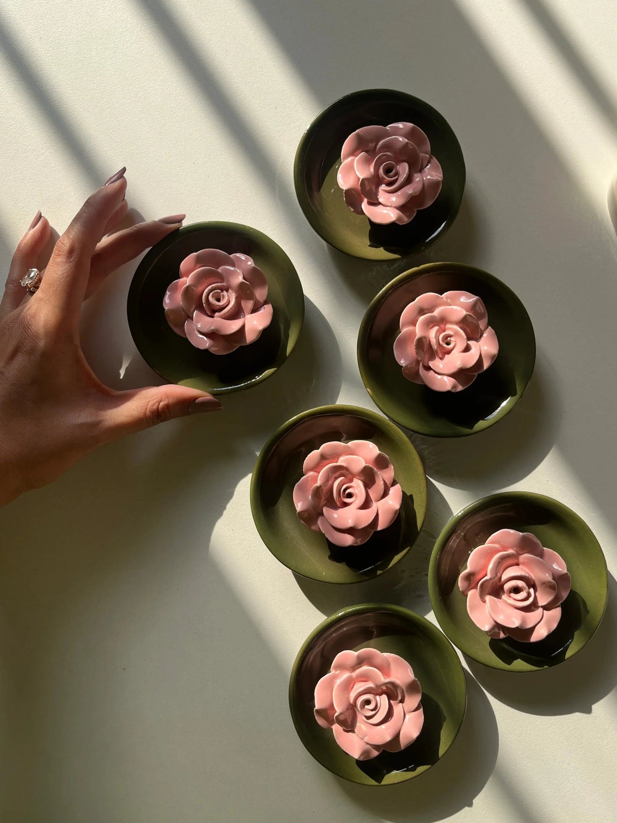 Six Posie incense stick holders with royal green base and pink flower, each holding a pink rose-shaped item, arranged on a white surface with strong diagonal shadows. A hand enters from the left, touching one of the dishes.