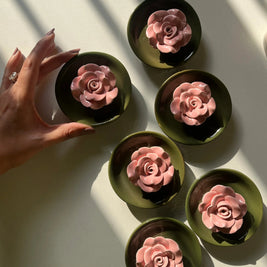 Six Posie incense stick holders with royal green base and pink flower, each holding a pink rose-shaped item, arranged on a white surface with strong diagonal shadows. A hand enters from the left, touching one of the dishes.