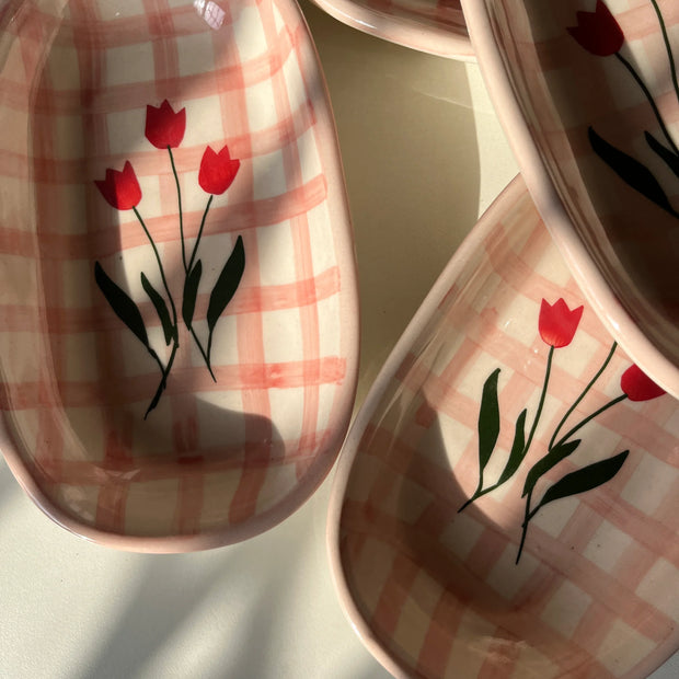 Three ceramic oval dishes with a pink and white gingham pattern and red tulip floral designs, arranged on a light surface with natural shadows.